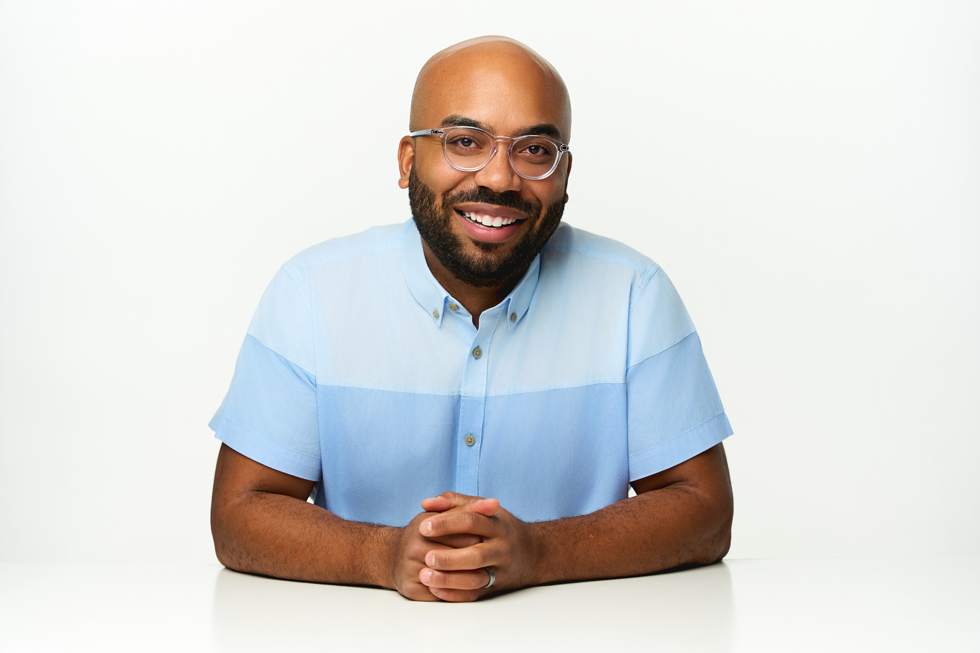 A photo portrait of Jason.  Jason is a bald, brown-skinned 30-something male with a neat, shortly cropped black beard, sitting at a white table.  He wears clear rimmed lawyerly glasses and a light blue two-toned collared short-sleeved dress shirt.  The photo is taken straight on and Jason is looking directly into the camera, in front of a white backgound.  His hands are folded and he's smiling.